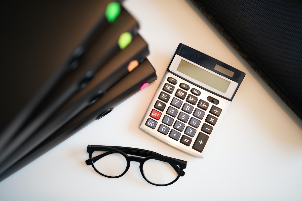 Objetos em uma mesa: uma calculadora, um óculos e 5 pastas pretas, uma em cima da outra